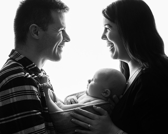 A black & white backlit semi-silhouette photo of parents with their newborn baby