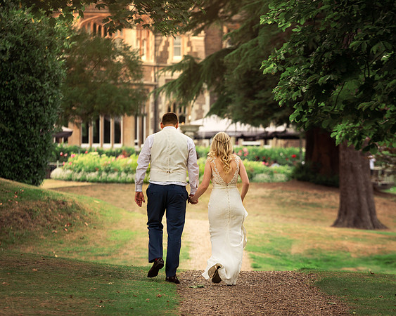 A wedding couple walk away from the camera in the shadow of Windsor's gothic Oakley Court hotel