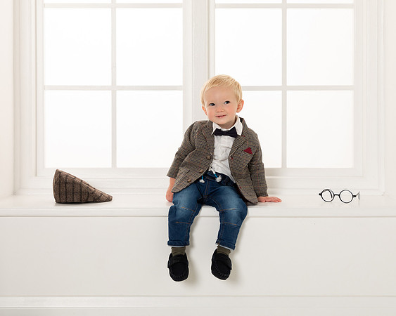 A toddler boy in jeans and jacket sits on a windsow sill on a fashion catalogue photoshoot