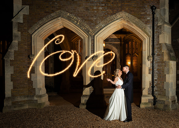 A wedding couple write the word 'love' with sparklers outside the gothic arches of Oakley Court in Windsor
