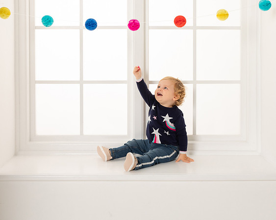 A catalogue images of a little girl sat by a window reaches up to a string of pom poms