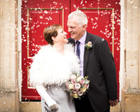 An older bride and groom smile at each other as confetti falls around them in front of Windsor Guildhall's red doors