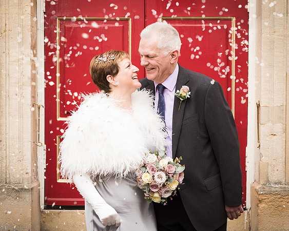 An older bride and groom smile at each other as confetti falls around them in front of Windsor Guildhall's red doors