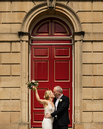 A bride raises her bouquet in the air while kissing her groom in front of the Windsor Guildhall's red doors