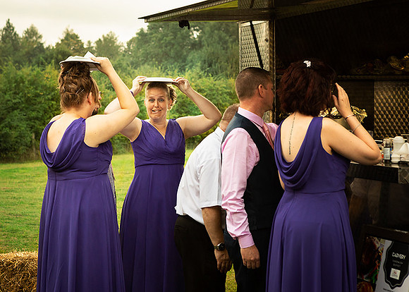 A bridesmaid makes a face whilst queueing for food and sheltering from the rain with a plate over her head