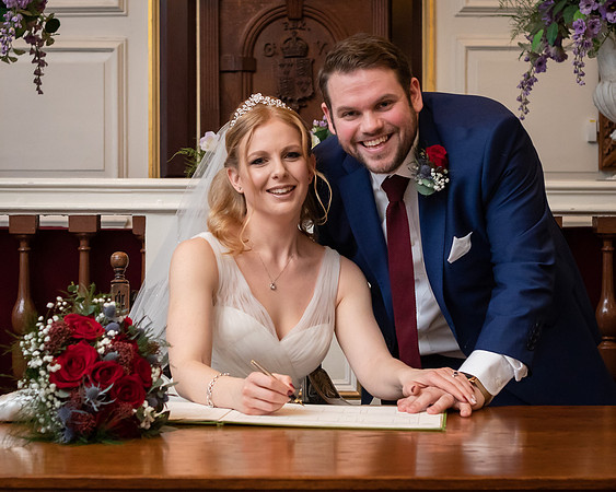A couple smile at the camera whilst signing the wedding register within Windsor Guildhall