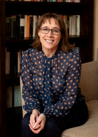 A woman sits on a sofa, relaxed smiling towards the camera with a bookshelf behind her