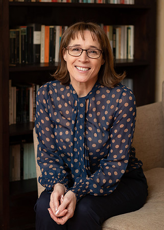 A woman sits on a sofa, relaxed smiling towards the camera with a bookshelf behind her
