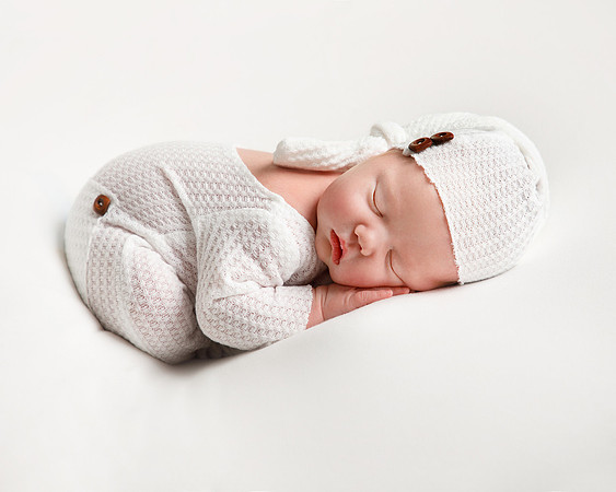 Newborn baby in a white sleepsuit and matching hat sleeps on her front