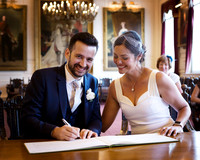 A wedding couple smile as they sign the register within Windsor Guildhall's Grand Chamber