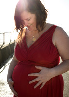 Pregnant mother in red dress smiles down at her baby bump while the sun shines over her