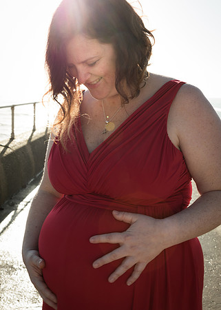 Pregnant mother in red dress smiles down at her baby bump while the sun shines over her