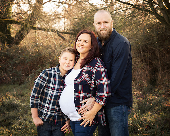 Family of three pose in an outdoor winter woodland, mum is expecting with a big baby bump