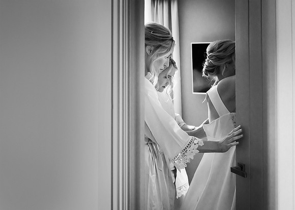 Through a doorway we glance a bride being buttoned into her wedding dress by her bridesmaids